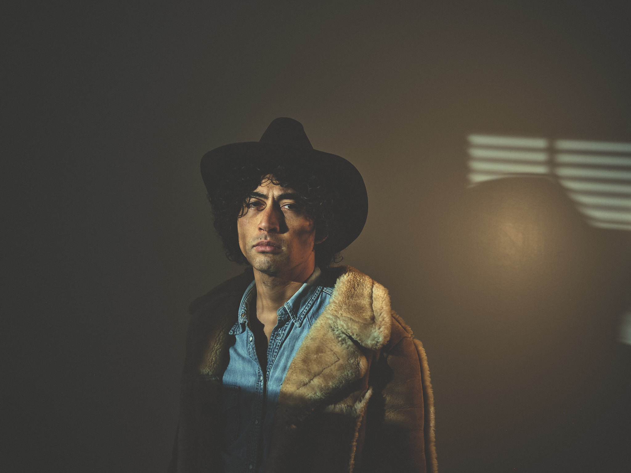 Cowboy in sheepskin jacket and hat, window blinds casting light across his face