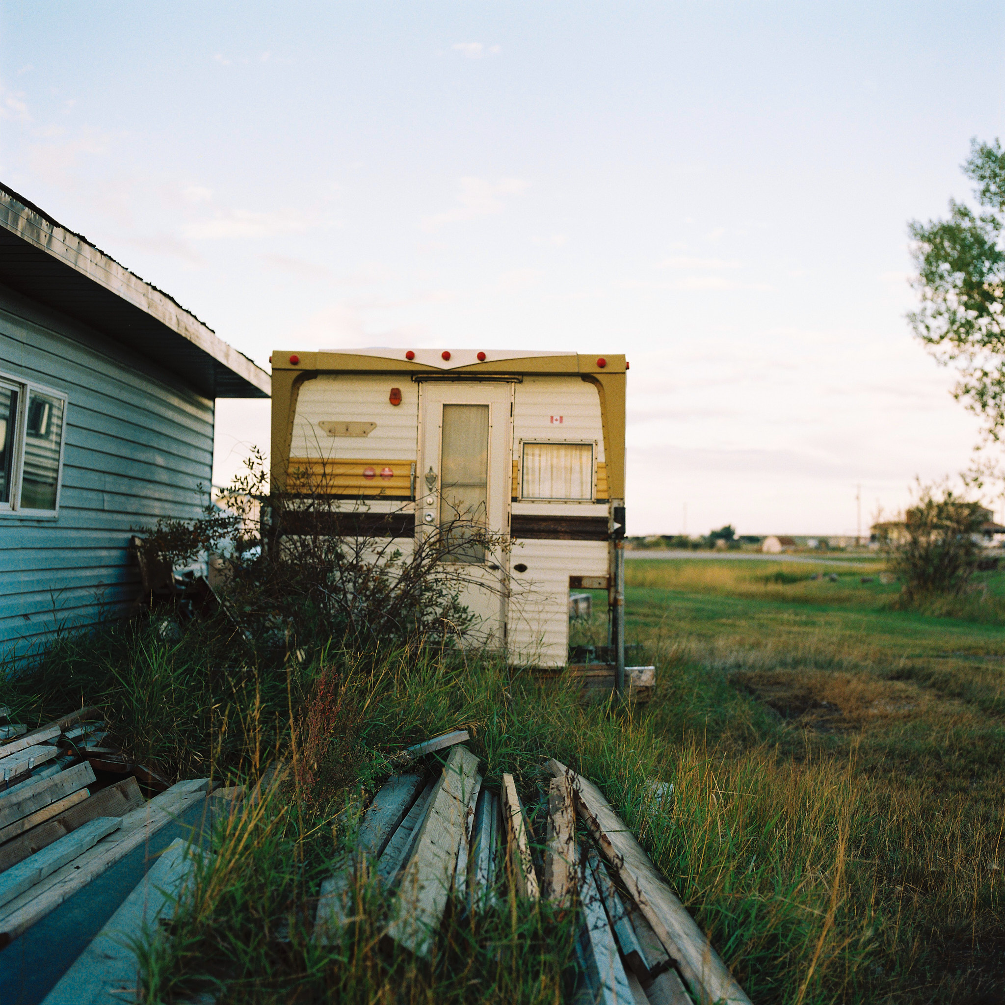 Abandoned camper trailer beside a blue house on the Alberta prairie, medium format